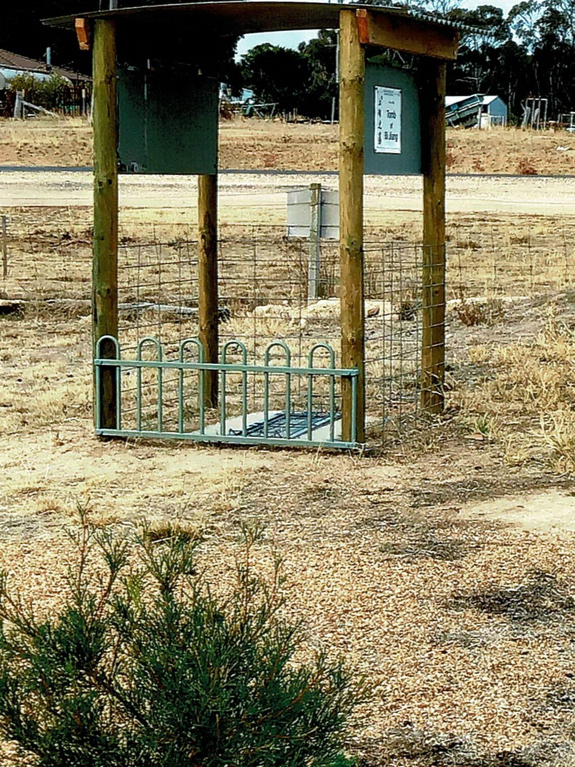 A gazebo and fence surrounding a headstone on the ground with roadworks and construction in the background.