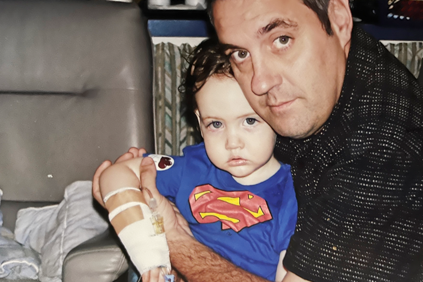 A young boy wearing a superhero outfit sits on a hospital bed with his dad