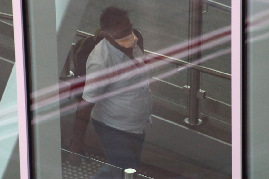 A man wearing a face mask and carrying a backpack walks in a corridor behind glass at Perth Airport.