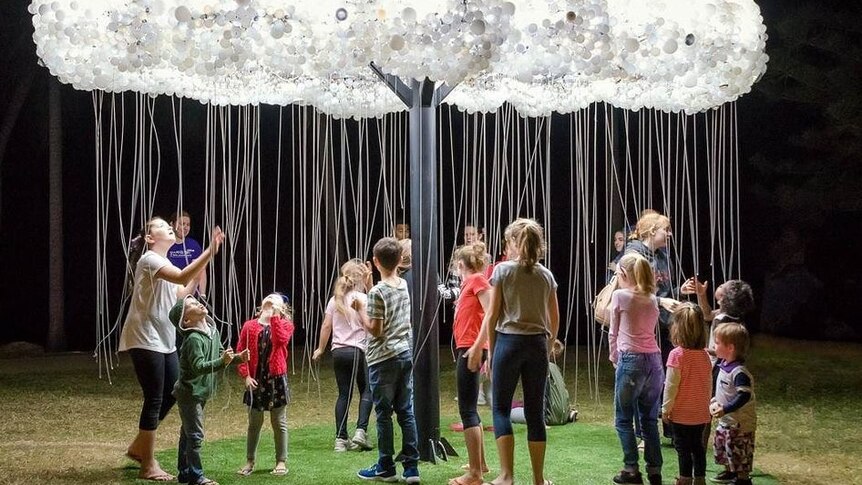 Children and adults underneath an artwork composed of a cloud made of lightbulbs and the cords that look like rain.