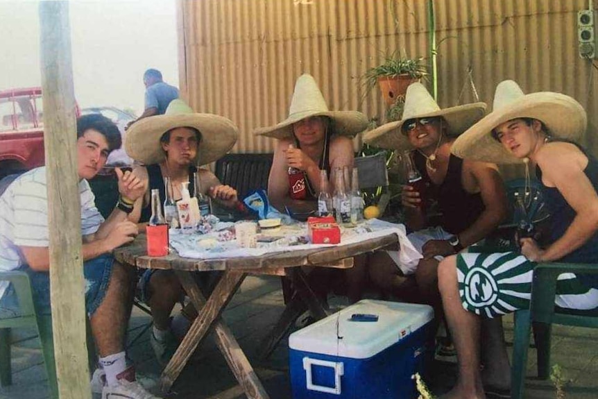 Five young men with Mexican hats drinking beers at a round picnic table, esky in front, corrugated yellow fence in background