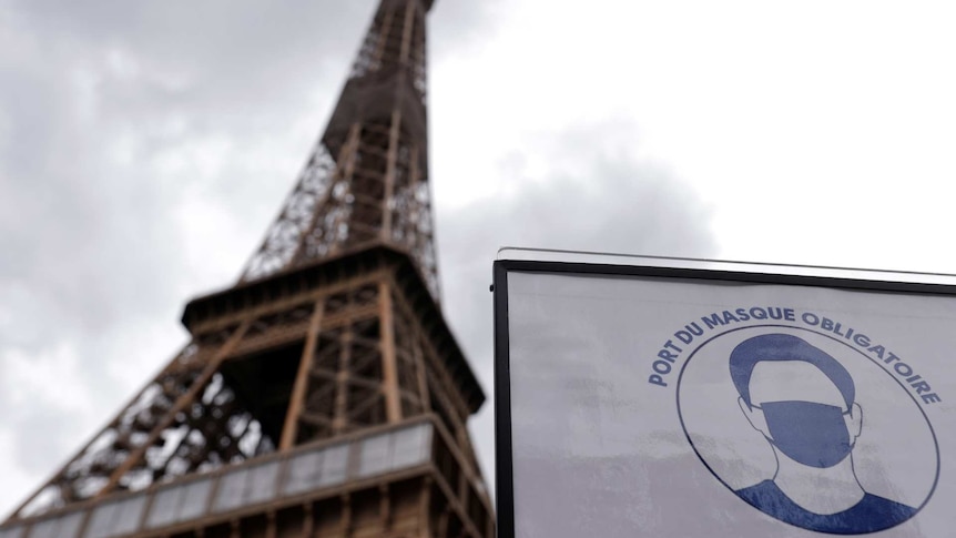 a sign warning about using face masks is shown in front of an out-of-focus the Eiffel Tower