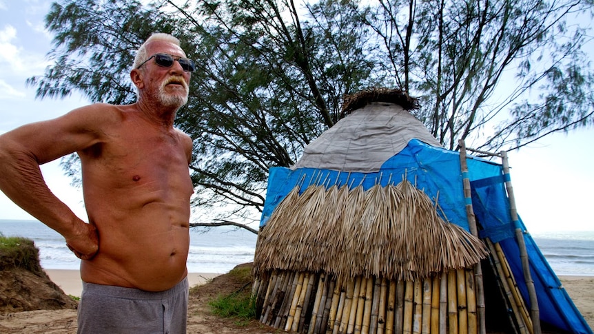 A man stands beside a hut made out of bamboo and tarpaulin, with the beach in background.