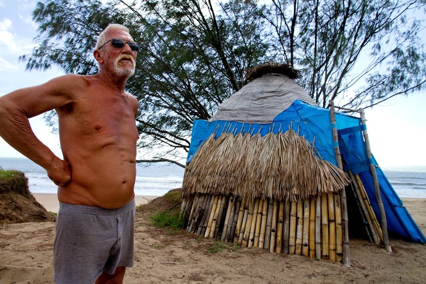 A man stands beside a hut made out of bamboo and tarpaulin, with the beach is in the background.
