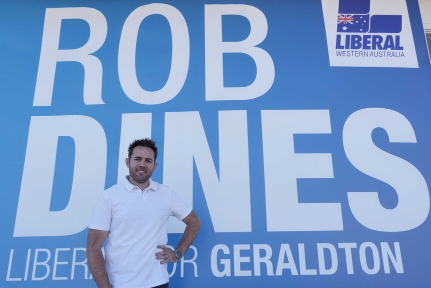 A man in white shirts stands in front of Liberal campaign sign with his name.