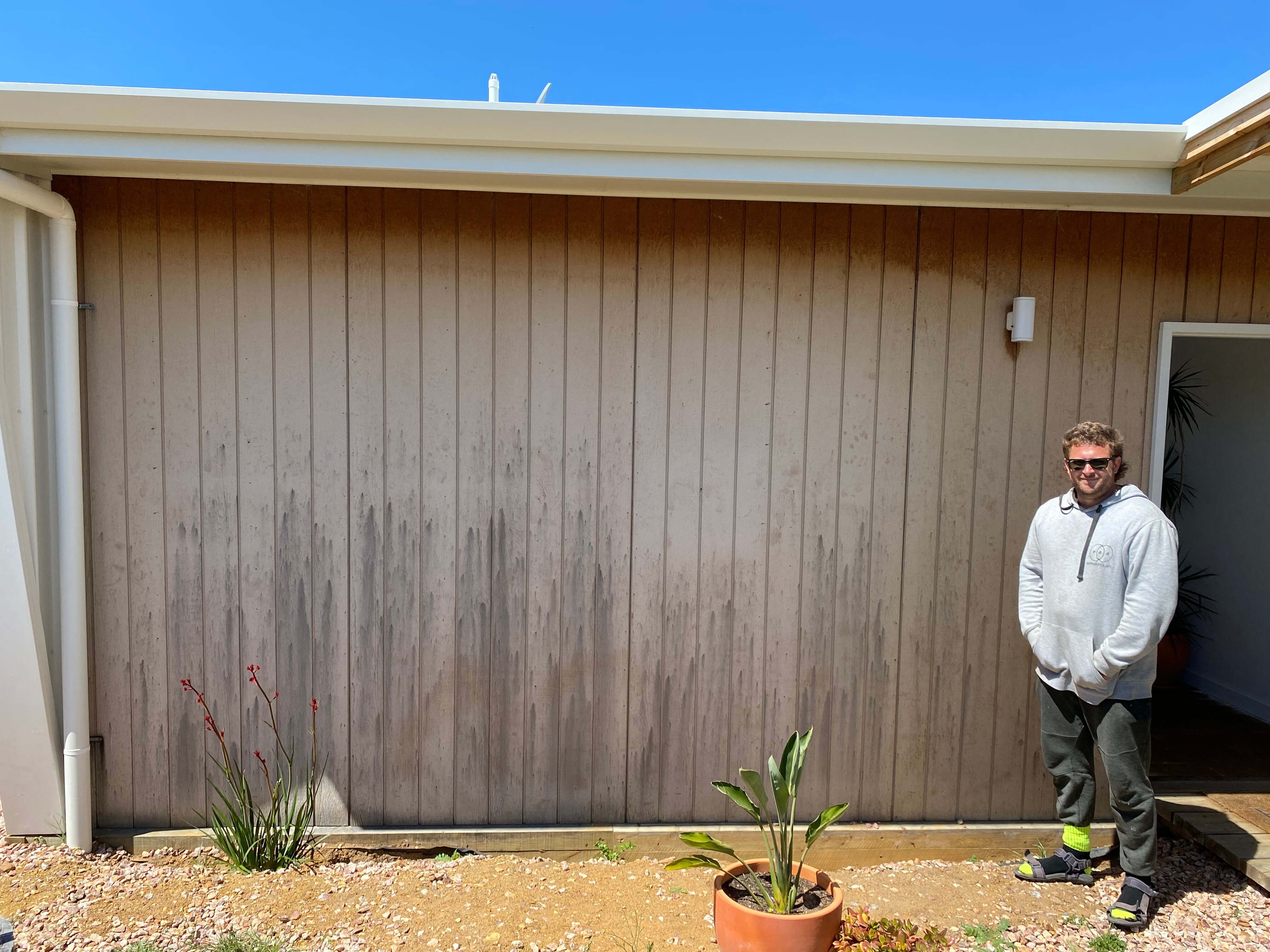 A man standing in front of the wall of a house with dark-coloured markings all over it.