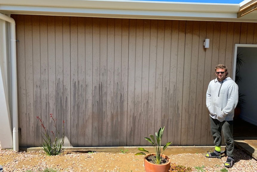 A man standing in front of the wall of a house with dark-coloured markings all over it.