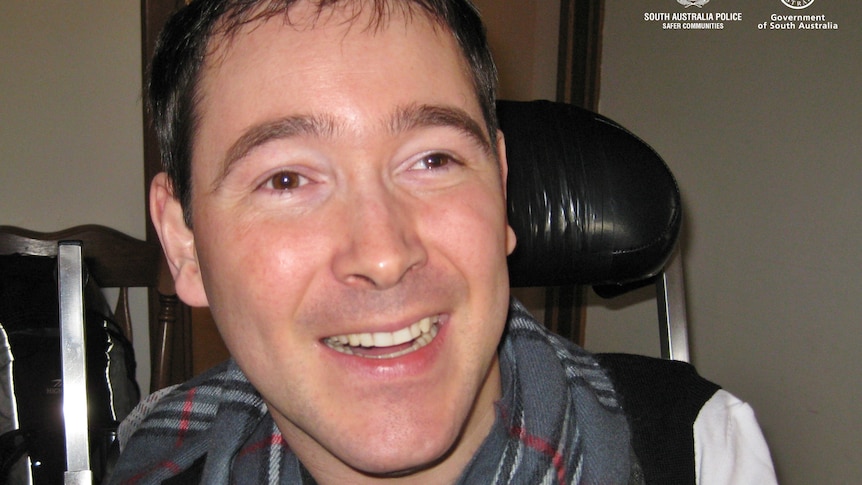 A man with dark hair wearing a grey checkered scarf smiles as he sits in a black wheelchair