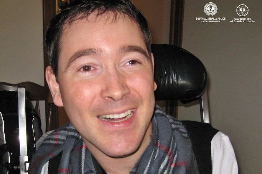 A man with dark hair wearing a grey checkered scarf smiles as he sits in a black wheelchair