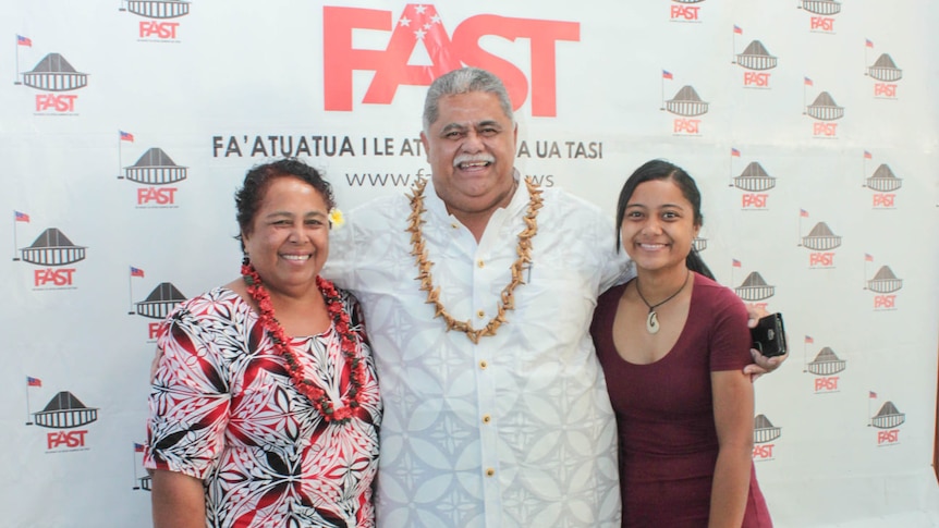 Two women and a man stand arm in arm and smile as they pose for a photo in front of a political party logo reading FAST