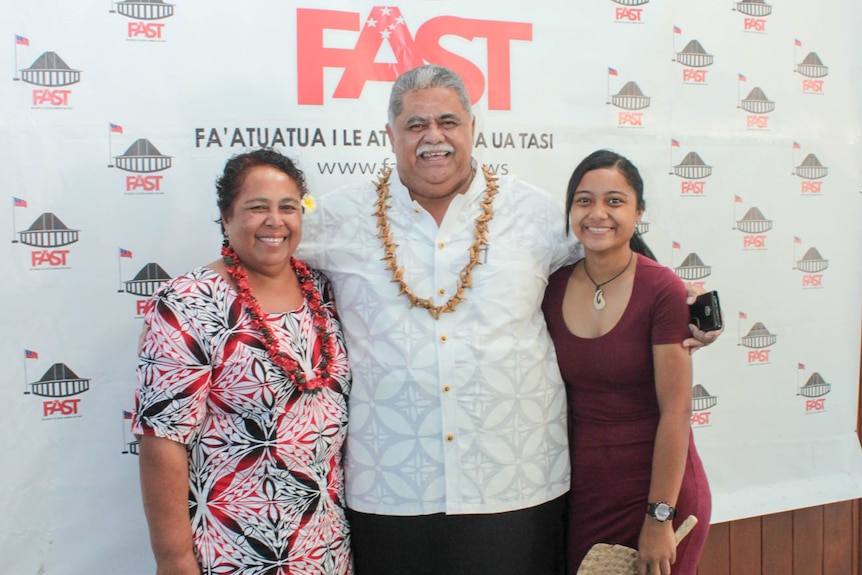 Two women and a man stand arm in arm and smile as they pose for a photo in front of a political party logo reading FAST