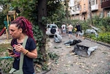 Woman with pink dreads stands in front of car wreckage 