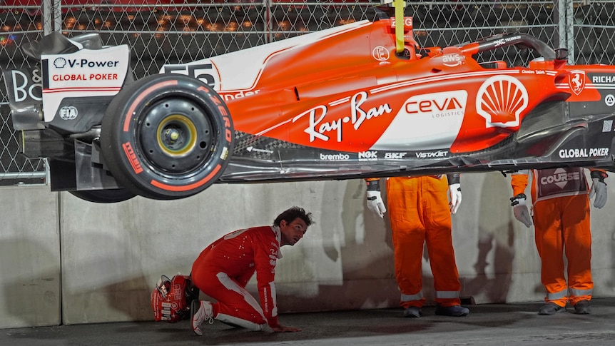 A Formula 1 driver crouches on the ground and looks up at the underside of his damaged car.
