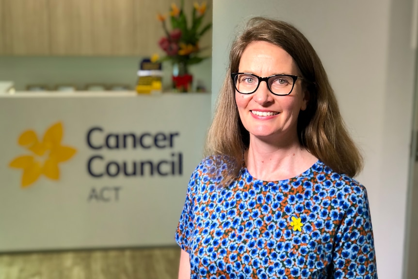 A woman smiles at the camera. In the background is a sign reading 'Cancer Council ACT'.