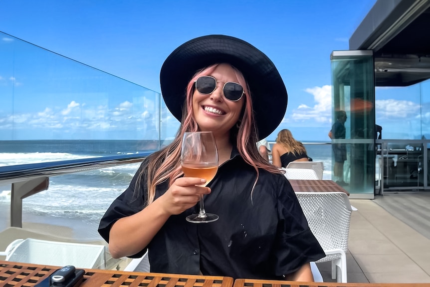 A smiling woman holds a glass of rose on an outdoor deck at the beach.