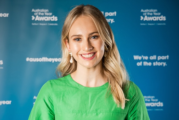 A smiling young woman with blonde hair, wearing a green blouse and holding a glass award trophy.