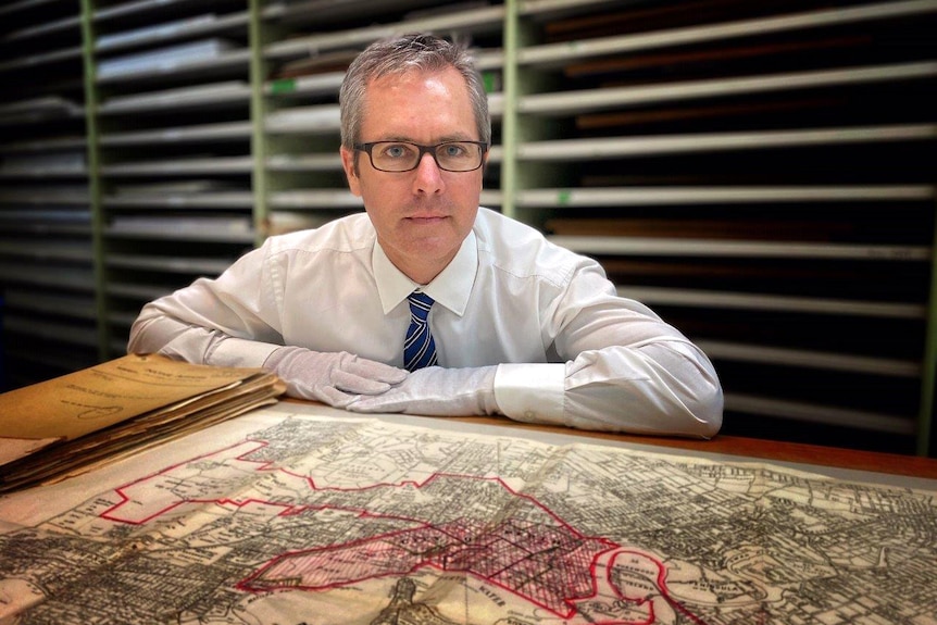 A man wearing glasses and gloves wits at a desk with a map in front of him and shelving behind him.