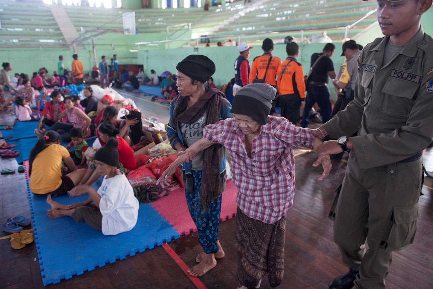 Evacuated people sit and stand in a gymnasium.