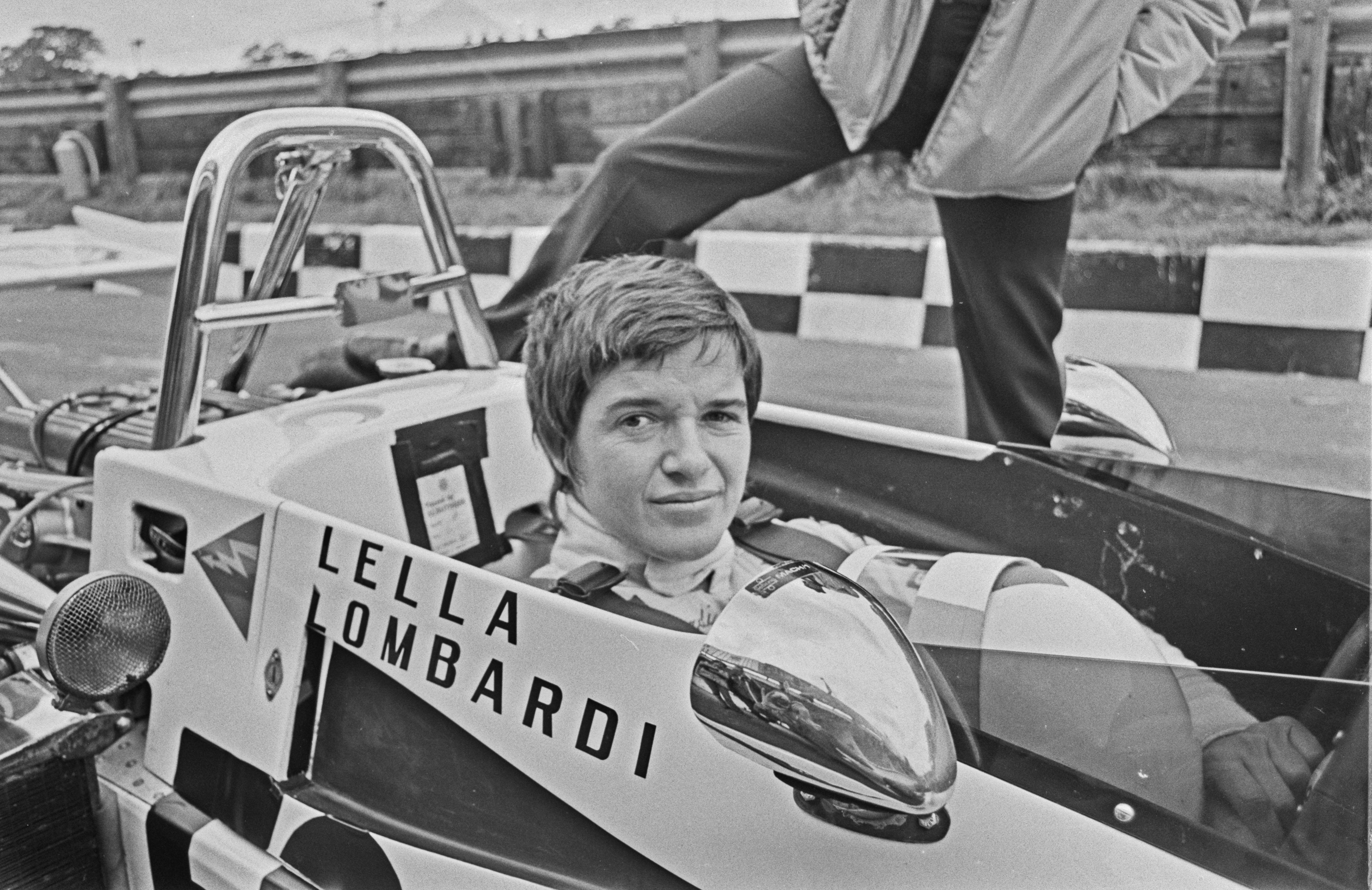 Uma foto em preto e branco de uma mulher com cabelo curto sentada em um velho carro de corrida aberto com "LELLA LOMBARDI" ao lado.