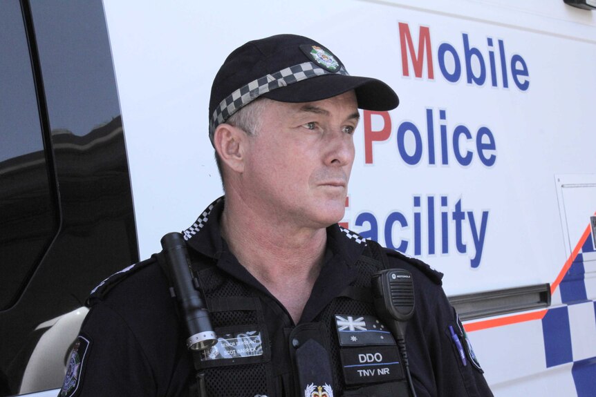 An older male police officer in uniform wearing a cap, standing in front of marked vehicle.