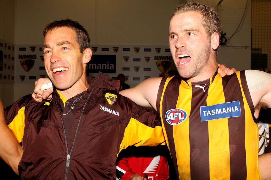 Hawthorn coach Alistair Clarkson stands with a beaming smile, singing, with his arm around the shoulder of one of his players.