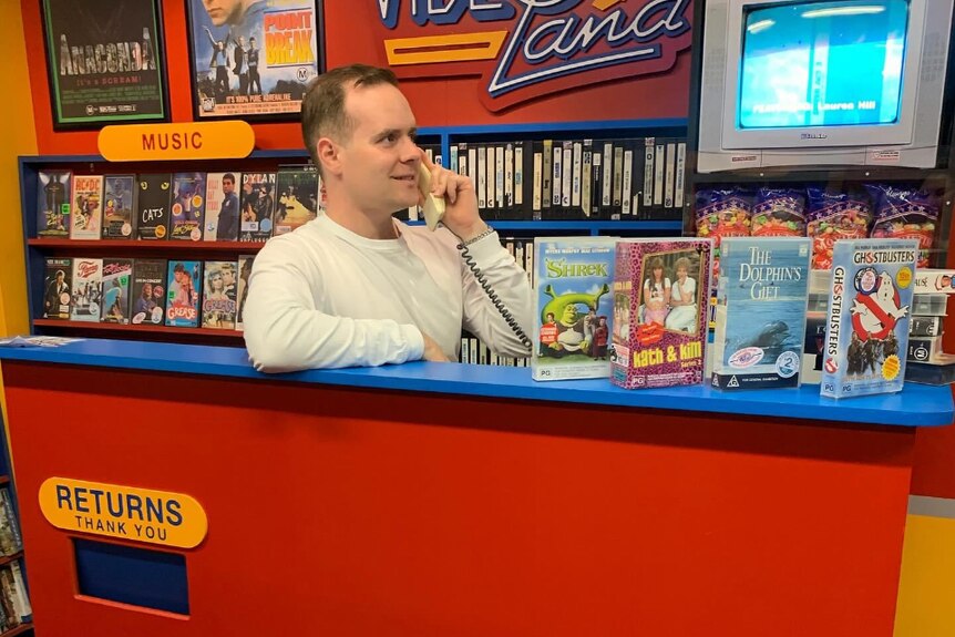 A man stands behind the counter of a film rental store, smiling while on the phone. 