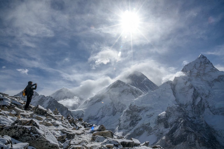 A person dressed in black clothes and wearing a backpack holds up a camera to take a photo of snow capped mountains