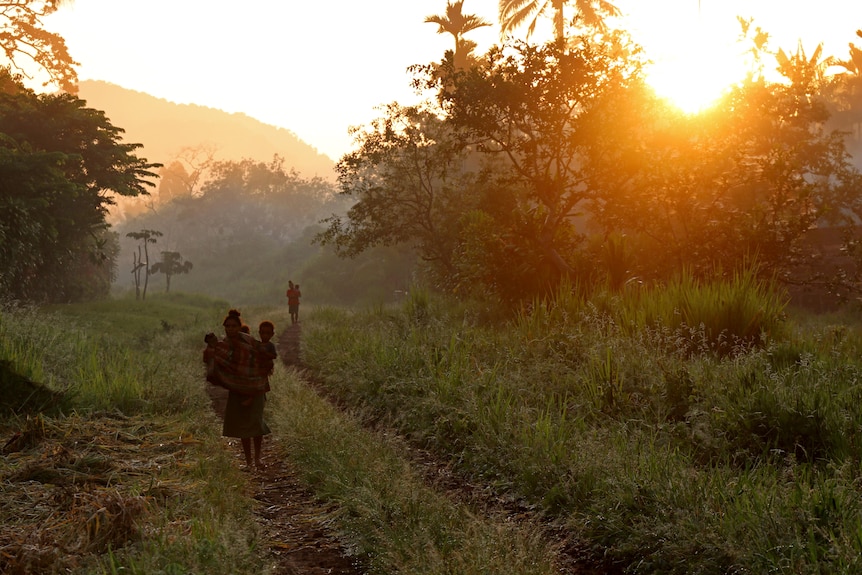 Two women walking down an overgrown dirt track carrying small children as the sun sets behind trees.