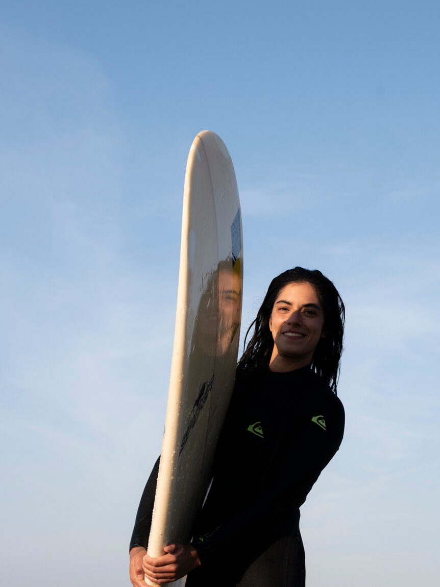 A mid shot of a woman holding a surfboard, smiling