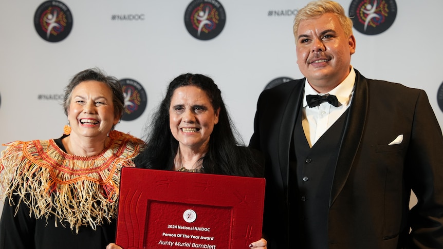 A middle-aged Indigenous woman holds up a red, framed award between two people in formal clothing in front of a white backdrop.