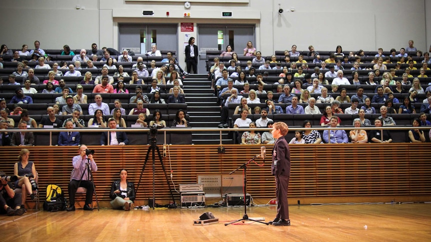 A primary school student standing at the microphone, in front of a packed auditorium.