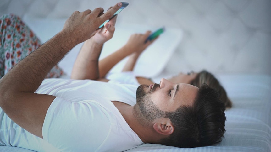 A man and a woman lie on a bed. Both are on their backs and are looking at their smartphones