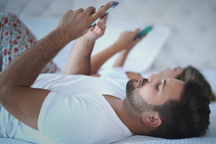 A man and a woman lie on a bed. Both are on their backs and are looking at their smartphones
