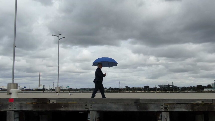 Alan Kohler walking along a jetty under cloudy skies holding an open umbrella