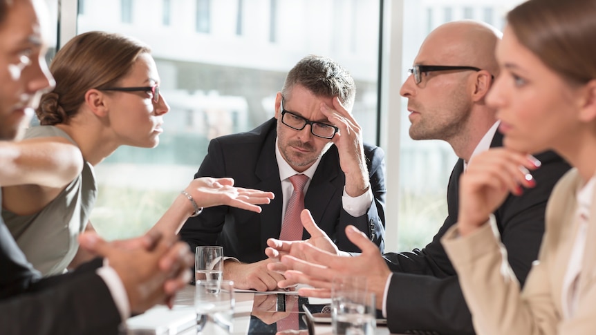 five people sit around a desk in a board room arguing