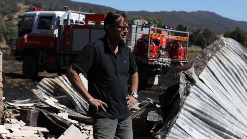 A man stands next to a burnt-down building. A fire engine is in the background.