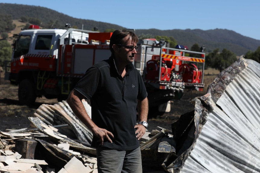 A man stands next to a burnt-down building. A fire engine is in the background.