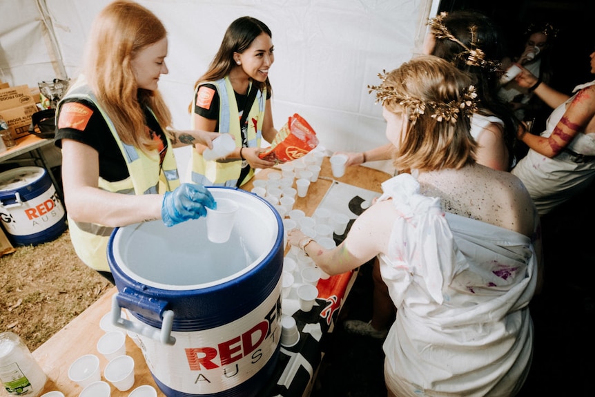 Two young women in high-vis vests offer cups of water to other young women wearing togas.