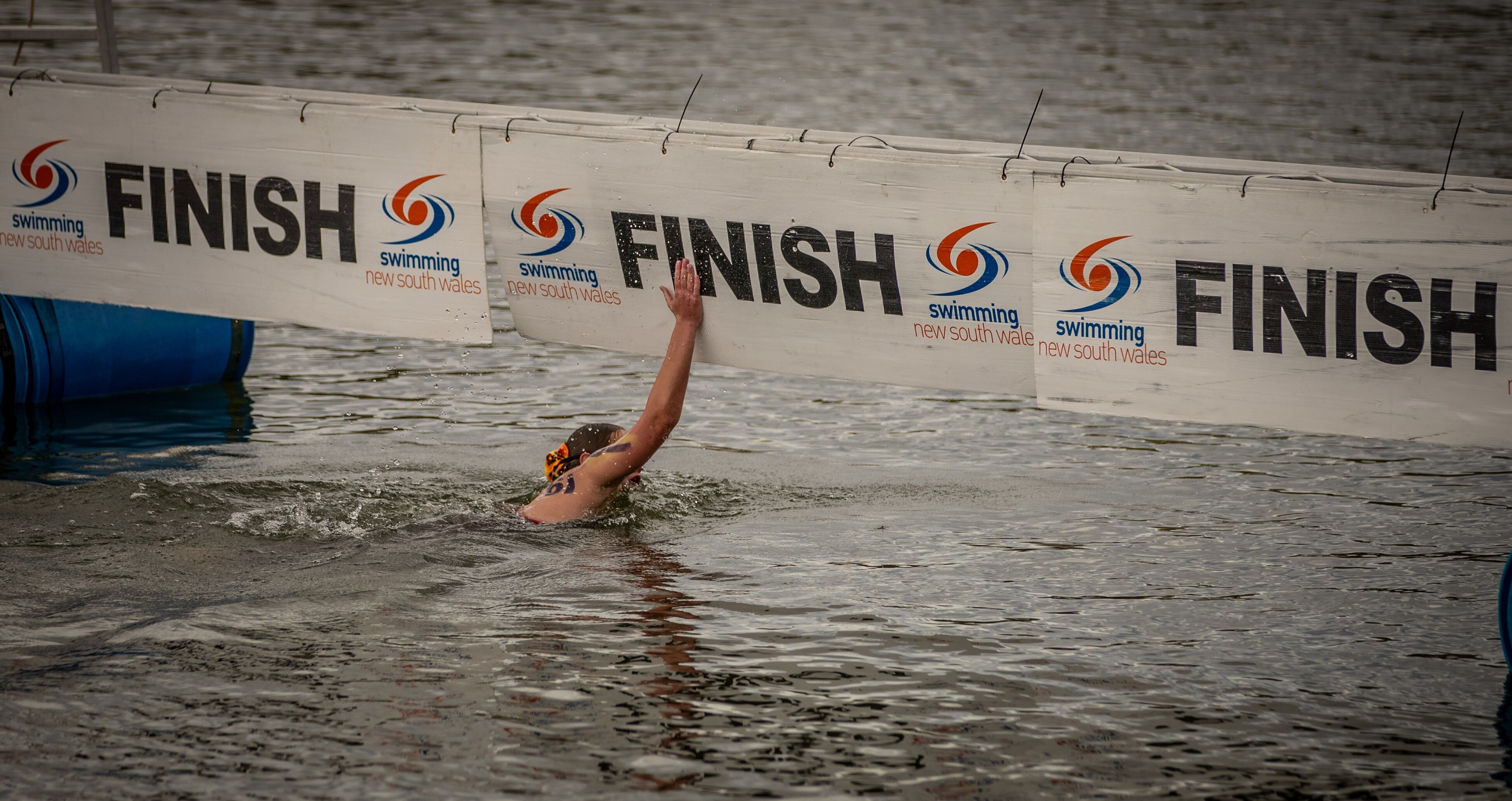 A swimmer in a body of open water slams her hand against a dangling finish line.
