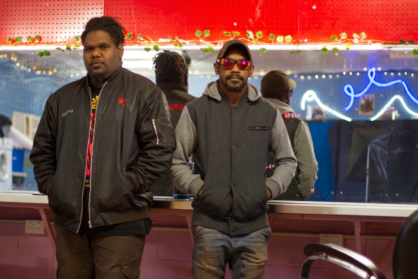 Two young men stand with their arms folded in front of a backstage mirror.