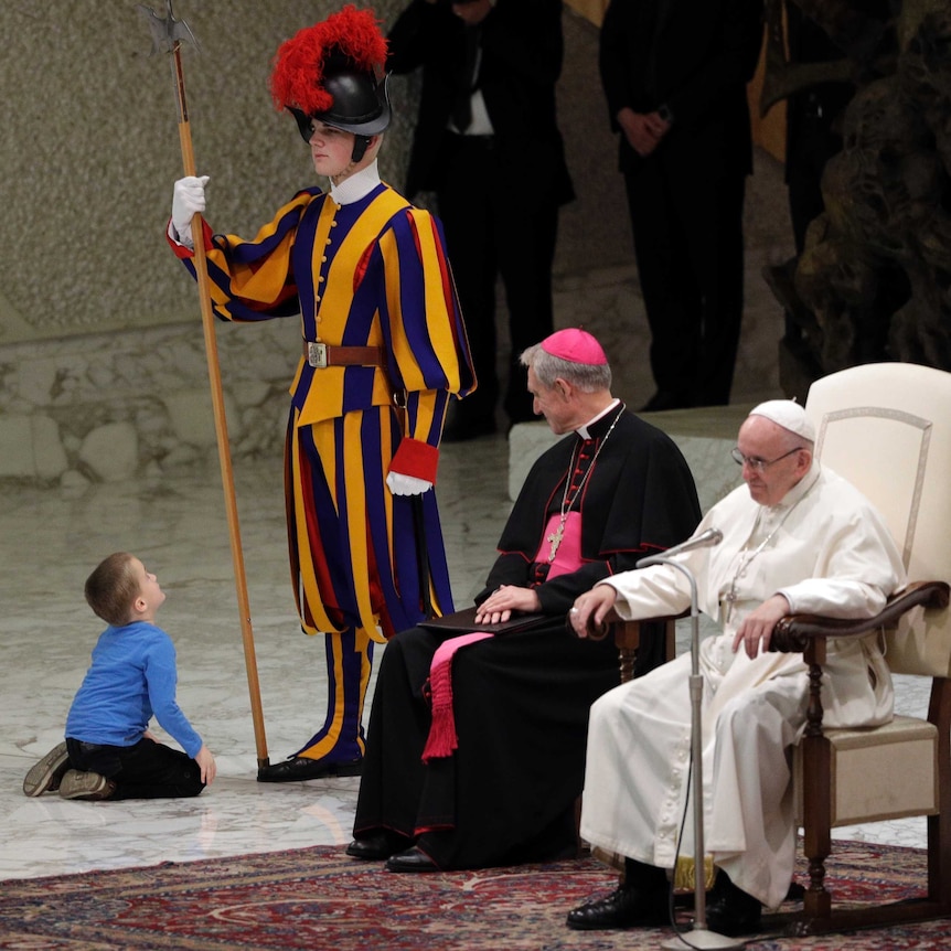 The young boy sits in front of the Swiss guard and looks up at him just metres away from the Pope on stage.