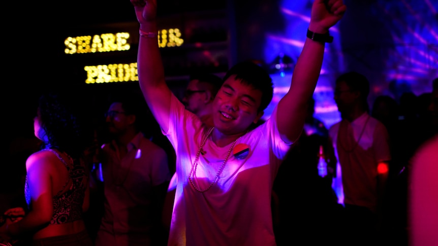 A man stands on a dancefloor with his hands in the air, bathed in a pink light. 