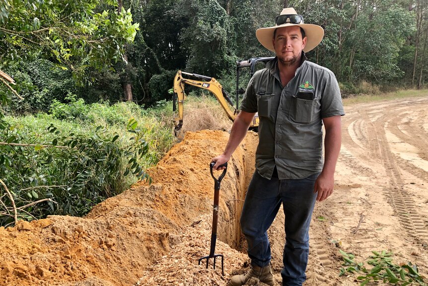 A farmer leans on a pitchfork in front of a trench partly filled with woodchips.