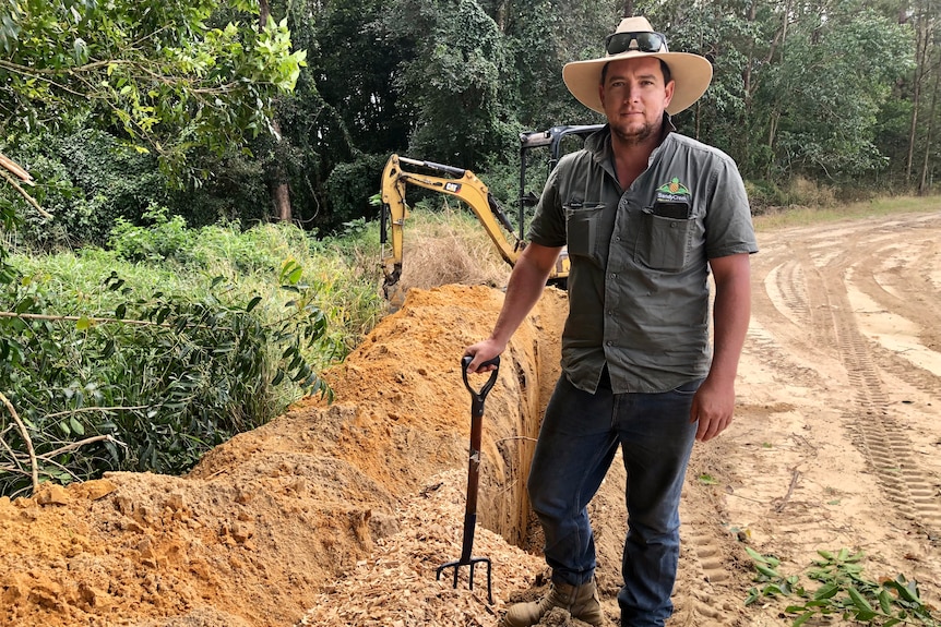 A farmer leans on a pitchfork in front of a trench partly filled with woodchips.