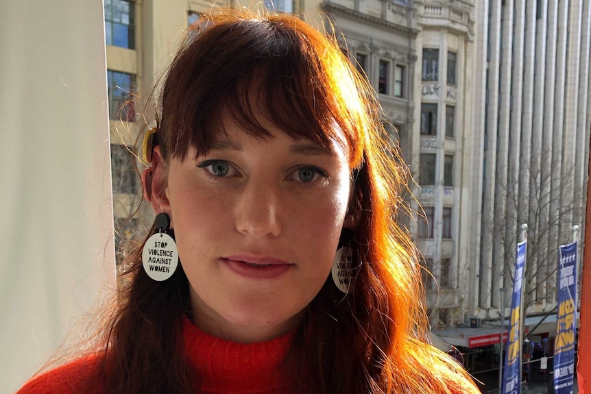A young woman with a red jumper and earrings that read 'stop violence against women'.