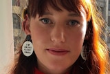 A young woman with a red jumper and earrings that read 'stop violence against women'.
