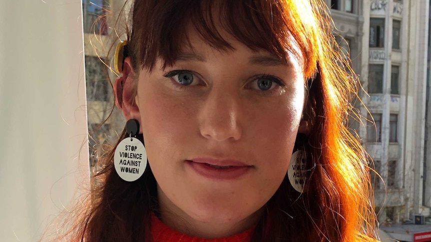 A young woman with a red jumper and earrings that read 'stop violence against women'.