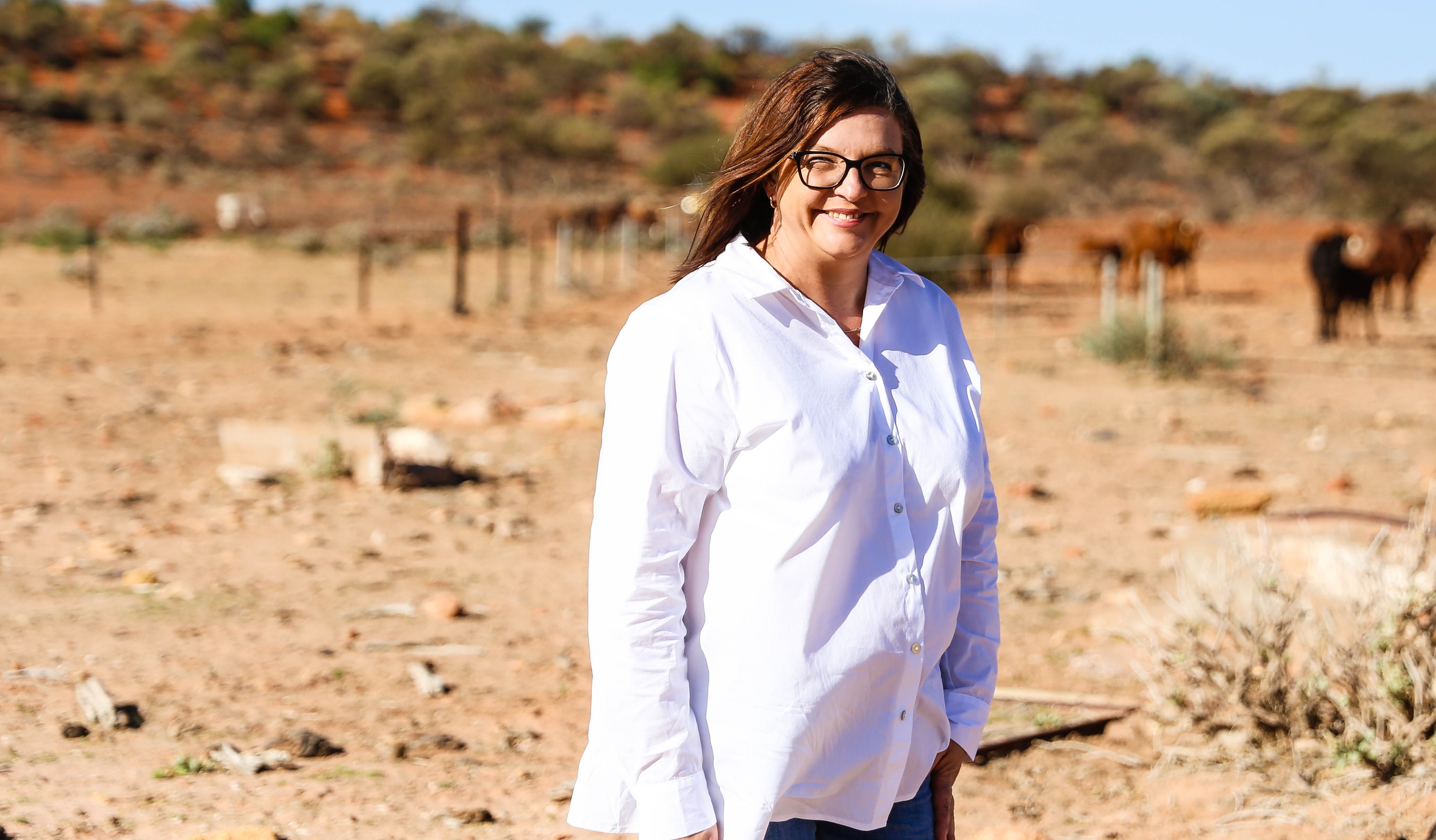 A woman in a white shirt smiles at the camera, fences and cows are in the background 