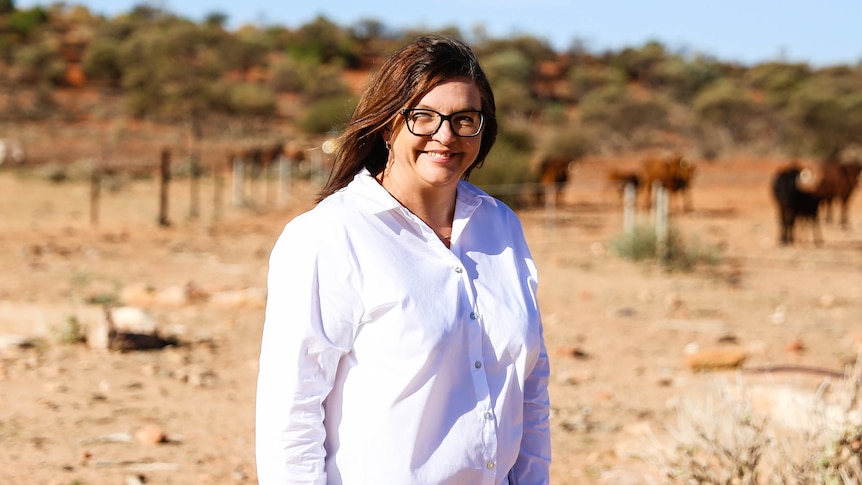 A woman in a white shirt smiles at the camera, fences and cows are in the background 