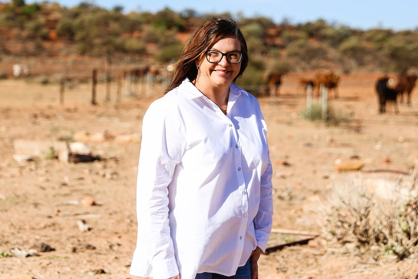 A woman in a white shirt smiles at the camera, fences and cows are in the background 